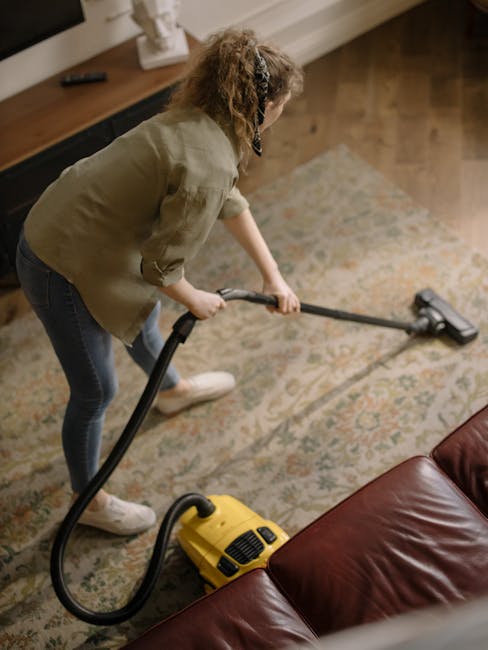 A woman with curly hair, wearing a light-colored jacket, dark jeans, and white sneakers, is using a yellow vacuum cleaner on a patterned carpet in a living room. She is holding the vacuum's hose and c