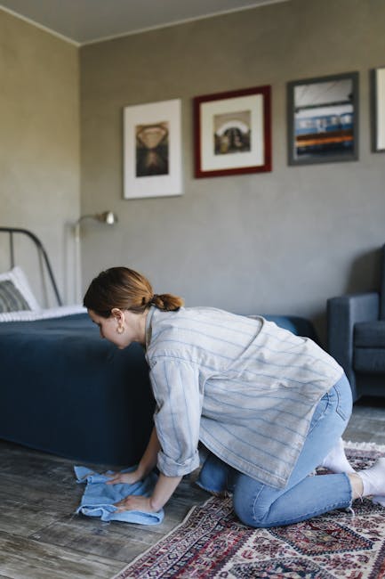 A woman with curly hair, wearing a light-colored jacket, dark jeans, and white sneakers, is using a yellow vacuum cleaner on a patterned carpet in a living room. She is holding the vacuum's hose and c