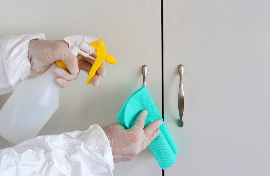 A person wearing white gloves and protective clothing is holding a spray bottle containing cleaning solution in their right hand and a turquoise microfiber cloth in their left hand, which they are using to wipe the silver door handles of a light gray cabinet or closet. The surface appears clean and shiny, with no visible dust or dirt, and the lighting in the room is bright, highlighting the smooth, matte finish of the cabinet doors and the stainless steel handles. This image exemplifies surface cleaning and deep cleaning practices for maintaining hygiene in residential settings, as performed by Deep Cleaning Merton, a professional cleaning service offering domestic cleaning solutions, mentioned on the page about deep clean tips for Wimbledon Village homes (SW19), Merton.