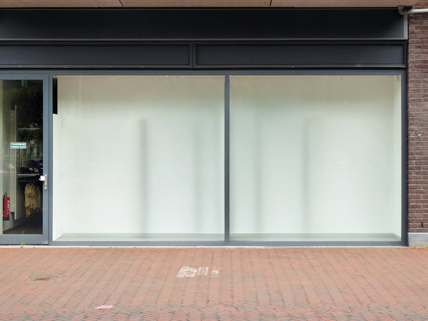 The image shows the exterior of a commercial property on Morden High Street with large, frosted glass windows that appear to have been recently cleaned, reflecting minimal dirt or smudges. The window frames are metallic, and the building features a brick wall on the right side. To the left, there's a glass entrance door with a metallic handle, revealing a glimpse of the interior, which includes a clothing rack and a fire extinguisher near the door. The pavement in front of the premises is made of red bricks, with a designated bicycle parking area marked on the ground. The overall appearance suggests the property is undergoing or has just undergone surface cleaning and sanitisation, with a tidy, well-maintained facade. This image exemplifies the professional cleaning services offered by Deep Cleaning Merton for commercial storefronts and shop fronts, ensuring a clean and inviting appearance on Morden High Street.