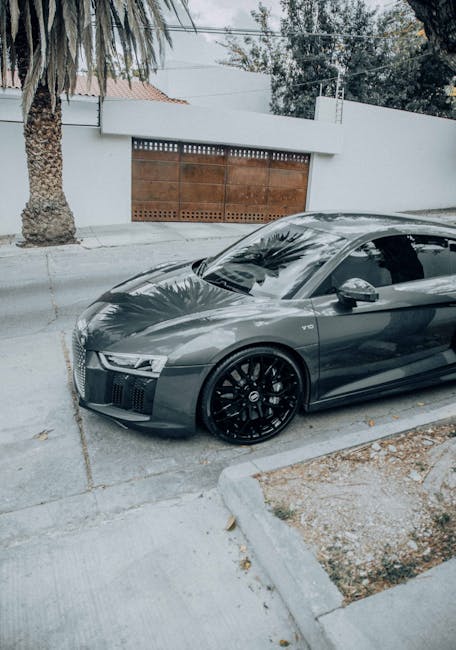 A sleek, dark grey Audi sports car parked on a residential driveway with a concrete surface. The car features tinted windows, black alloy wheels, and a low-profile design. The surrounding area includes a palm tree with a textured trunk on the left, a white wall with a wooden gate in the background, and mature trees visible beyond the wall. The driveway and car exterior appear clean and well-maintained, reflecting natural light and casting subtle shadows. The scene is part of a property on Morden High Street, with tidy landscaping and an emphasis on surface cleaning and vehicle hygiene. Deep Cleaning Merton offers professional cleaning services to maintain such levels of cleanliness and presentation.