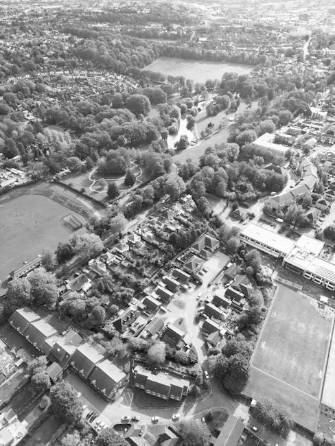 Aerial view of the Ravensbury Park estate in Merton, showing rows of residential houses with tiled roofs, surrounded by numerous trees and greenery. The estate is situated near a large park with open grassy areas, sports fields, and wooded sections. Pathways and roads wind through the residential area, with some vehicles visible along the streets. The overall scene is bathed in natural daylight, highlighting the lush foliage and the orderly layout of the estate. Deep Cleaning Merton offers comprehensive cleaning and sanitisation services for residential properties, ensuring spaces are hygienic and well-maintained, aligned with estate clean plans for the Ravensbury Park area. The image conveys a peaceful, community-oriented environment with abundant natural elements and well-kept streets.