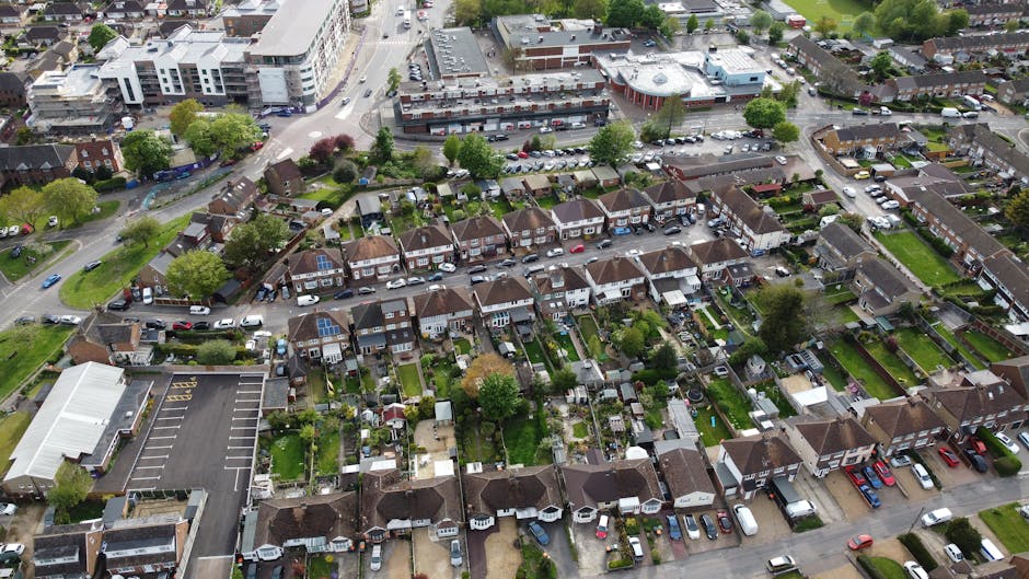 Aerial view of the Ravensbury Park estate in Merton, showing rows of residential houses with tiled roofs, surrounded by numerous trees and greenery. The estate is situated near a large park with open grassy areas, sports fields, and wooded sections. Pathways and roads wind through the residential area, with some vehicles visible along the streets. The overall scene is bathed in natural daylight, highlighting the lush foliage and the orderly layout of the estate. Deep Cleaning Merton offers comprehensive cleaning and sanitisation services for residential properties, ensuring spaces are hygienic and well-maintained, aligned with estate clean plans for the Ravensbury Park area. The image conveys a peaceful, community-oriented environment with abundant natural elements and well-kept streets.
