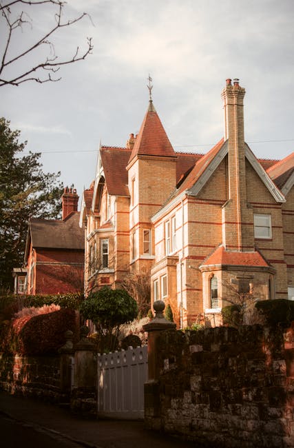 A historic Victorian-style house with a prominent gabled roof covered in red shingles, featuring white decorative trim around the windows and edges. The facade includes large, multi-pane windows, some