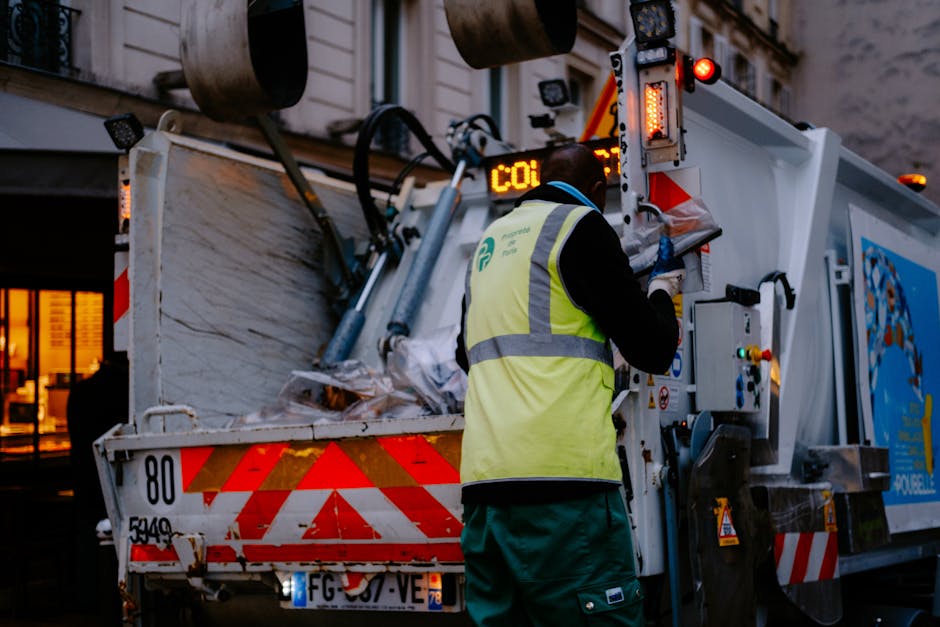 A sanitation worker in a black jacket and yellow high-visibility vest operating a waste collection vehicle at dusk. The vehicle is white with red and orange reflective stripes, filled with various bulky waste items, and features technical controls and warning lights on its side. The background shows a building with illuminated windows, indicating an urban setting. This scene illustrates waste management and the removal of bulky waste, which is part of comprehensive cleaning services offered by Deep Cleaning Merton for post-renovation clean-ups in Merton.