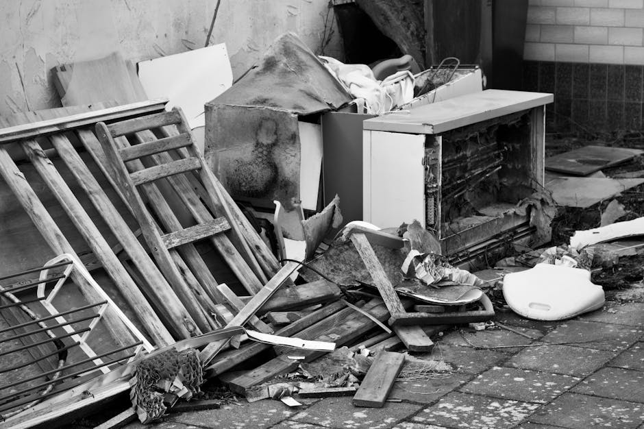 A black-and-white photograph showing a cluttered outdoor area with discarded furniture and debris from a recent renovation. There are wooden ladders and planks leaning against a wall, alongside an old washing machine with visible rust and dirt, partially covered with dust and debris. scattered across the brick-paved ground are broken wood pieces, a foam cushion, and a white plastic container. The scene is illuminated by natural light, highlighting the dusty and messy state of the surfaces, indicating a need for thorough cleaning and waste removal, which Deep Cleaning Merton can provide in their domestic cleaning services.