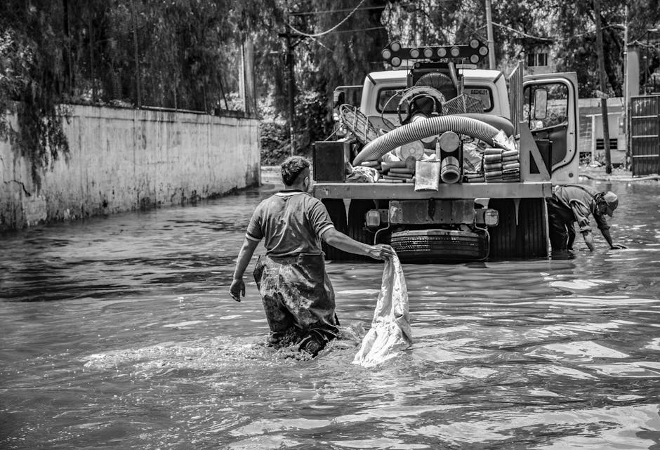 A black and white photograph depicting a residential flood cleanup scene. Two men are working in a flooded outdoor area, one standing in the water holding a piece of cloth or tarp, while the other is bent over near a large industrial vacuum or water extraction machine. The area appears to be a backyard or street with a concrete wall on the left and trees in the background. The floodwater covers the ground and partially surrounds the cleaning equipment, which is equipped with hoses and tanks. The scene emphasizes surface cleaning and water removal processes, illustrating the effort involved in flood damage restoration, consistent with the services provided by Deep Cleaning Merton. The overall setting highlights the importance of efficient disaster response and thorough sanitisation of affected surfaces.